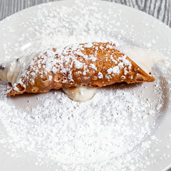 A Tony Sopranos cannoli, filled with cream and dusted with powdered sugar, on a white plate.