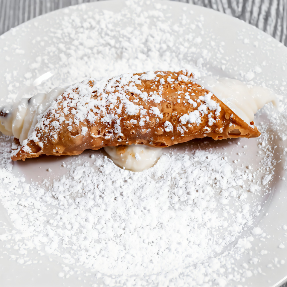 A Tony Sopranos cannoli, filled with cream and dusted with powdered sugar, on a white plate.