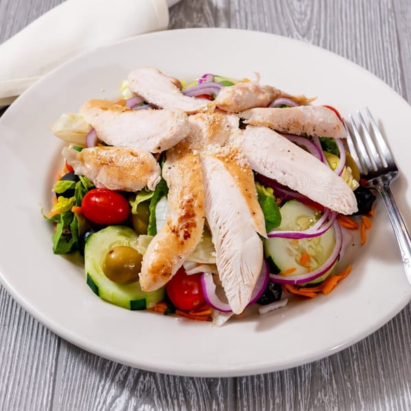 Grilled chicken salad with lettuce, tomatoes, cucumbers, red onions, and olives on a white plate.