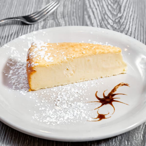 Slice of cheesecake with powdered sugar and chocolate drizzle on a white plate.